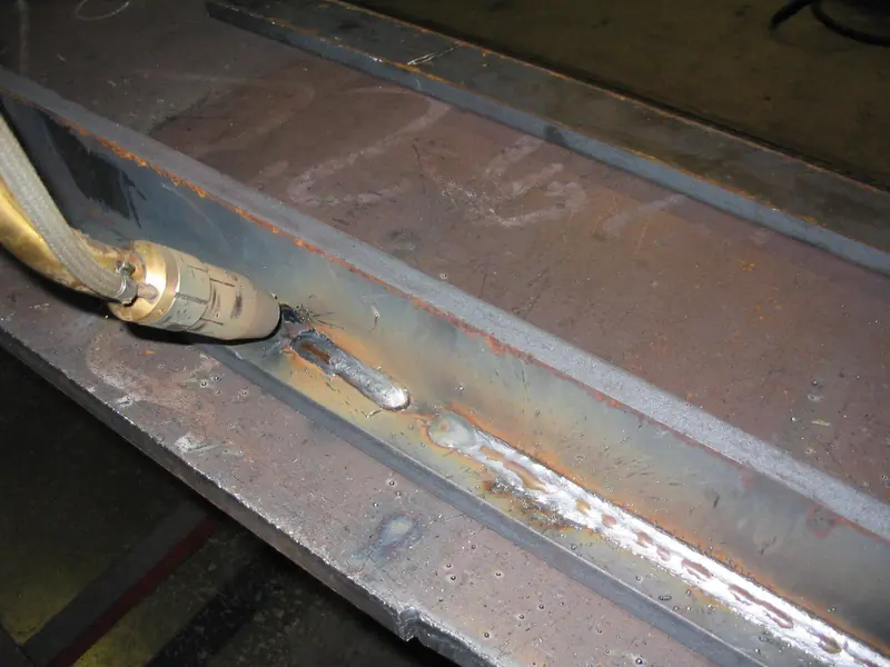 A close-up of a continuous arc-welded seam along a steel beam, with the ripple pattern of the weld bead in focus.